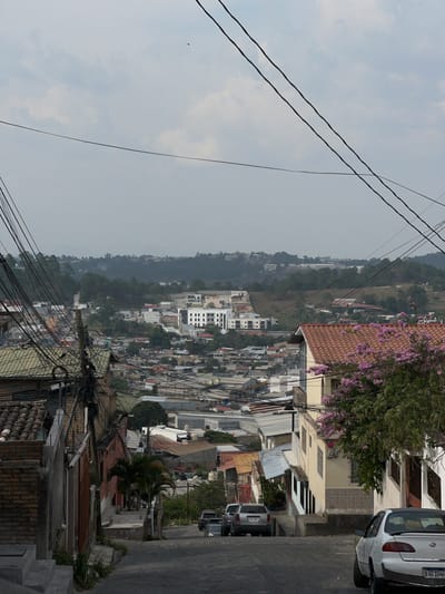 The image shows a cityscape in Santa Rosa de Copán, Honduras. It is taken from an elevated position looking down at buildings and houses of varying sizes and designs. A valley stretches out in the background under an overcast sky. Overhead power lines crisscross the foreground of the image. Along the right side of the frame is a street with parked cars, including a white Toyota with license plate HAD 1046. Vegetation, including trees with pink flowers, is visible. The general atmosphere is one of urban density and a somewhat hazy environment.