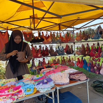 An outdoor market stall in Kebondalem, Indonesia, is depicted under a large yellow canopy. A woman wearing a black hijab and a brown abaya is shown interacting with a small brown purse. The stall displays a variety of merchandise, including numerous bras of various colors and styles hanging from racks, as well as folded items of clothing, possibly underwear or children's clothing, piled on tables. The background reveals a rural setting with greenery and water, suggestive of rice paddies. The lighting indicates it is daytime.