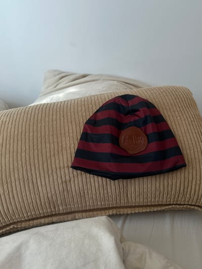 A red and black striped hat with a brown leather logo patch is lying on a beige pillow. The location is not discernible from the image.