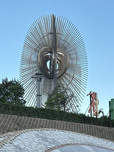 The image shows a metal sculpture of a stylized flower or sunburst. The sculpture is made of concentric rings of metal rods radiating outward. There is also a sculpture of a horse and rider that is in proximity. The sky is blue. The location is Taihe District, China.