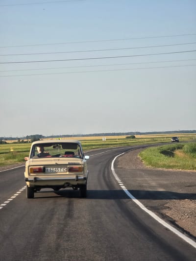The image shows the rear view of a light-colored car, likely a Lada, driving on a paved road in a rural area. The car has a license plate that reads "А 3385 ГС". Inside the car, one can see two people. The road is bordered by grassy fields and some trees in the distance, suggesting a countryside setting. The sky is clear and light, indicating it's daytime, possibly late afternoon or early morning due to the long shadows. The overall mood is serene and peaceful, capturing a moment of travel through a quiet landscape. The scene appears to be set in Ровенскаслабодскі сельскі Савет, Belarus.