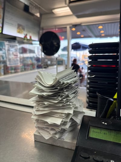 A close-up, eye-level shot captures a stack of receipts held together by a thin, pointed object, likely a pen or pencil, standing upright on a metal counter. The receipts are a mix of white and light-colored paper, folded and layered, creating a dense, irregular pile. To the right of the receipts is a black electronic device with a green illuminated screen displaying "Guest #:" followed by "101547". The device has several buttons and a textured grip area. In the background, through a glass partition, a blurred scene of a business interior is visible. This includes a menu board on the left, shelves with various items, and a doorway leading outside to a bright, slightly hazy environment with blurred figures and awnings. The overall setting appears to be a point-of-sale area in a commercial establishment.