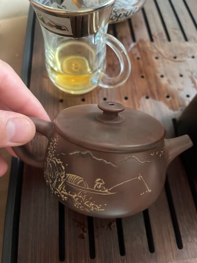 A close-up shot captures a person's hand gently holding the handle of a small, brown teapot with intricate carvings. The teapot sits on a dark, wooden tea tray with a slatted surface. In the background, a clear glass cup filled with amber-colored liquid, presumably tea, is visible, resting on the same tray. The scene appears to be an indoor setting, possibly a home or a tea house, with soft lighting. The overall impression is one of a quiet, contemplative moment focused on a tea ceremony. The specific location in Saransk, Russia, cannot be determined from the visual information provided in the image.