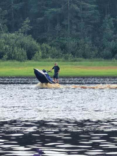 A man rides a jet ski on a body of water, kicking up a brown spray. He is wearing a dark short-sleeved shirt with a blue pattern and blue patterned shorts. The jet ski is dark blue and black with a prominent blue stripe. In the background, a dense green forest lines the edge of the water, with a strip of grassy field between the water and the trees. The sky appears overcast, suggesting it might be a cloudy day or late afternoon. The overall scene depicts a recreational activity on the water with a natural, wooded backdrop. There is no visible text in the image.