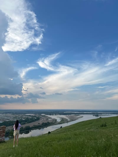 The image shows a woman standing on a grassy hill overlooking a wide river and a floodplain. The woman is facing away from the camera, wearing a light purple top and light-colored shorts. Her long, dark hair is visible. The hill is covered in green grass and wildflowers. In the distance, the river winds through a landscape of trees and fields, with buildings and structures visible on the horizon. The sky is a mix of blue with dramatic clouds, some of which are white and wispy, while others are darker and more voluminous. A ruined brick structure is partially visible on the left side of the frame, near the woman. The scene is likely set in a natural landscape near Britvino, Russia, given the presence of a river and floodplain.