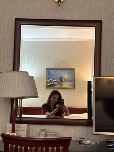 A woman is taking a mirror selfie in a room. She is holding a black smartphone in her right hand, angled towards herself. She is wearing a brown v-neck shirt. In the reflection of the mirror, a bed with white bedding is visible behind her. To the left of the mirror, a cream-colored lampshade with a gold trim is visible. Above the bed, a framed painting of a windmill by a body of water is hanging on the wall. To the right of the painting, a portion of a black television screen is visible. The room appears to be a hotel room, with textured beige wallpaper on the walls. A wooden desk is in the foreground, with a dark wooden chair partially visible. There is a small white sign with text in Russian on the desk. The overall lighting is soft and warm. The location is a hotel room in the city of Zhuralyovo (Kemerovo Oblast), Russia.