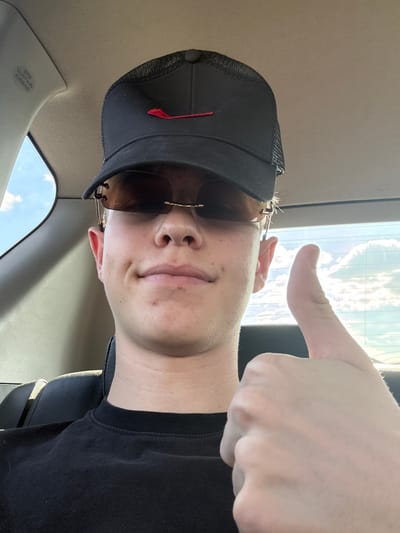 The image shows a close-up, sideways selfie of a young person in a car. The person is wearing a black trucker hat with a red logo and brown-tinted sunglasses. Their face is partially visible, with a slight smile. A hand is raised in the foreground, giving a thumbs-up gesture. The background appears to be the car's interior, with a window showing a bright, cloudy sky, suggesting it is daytime. The lighting indicates sunny weather. Visible text on the car's interior trim reads "SRS CURTAIN AIRBAG". The overall mood is casual and positive.