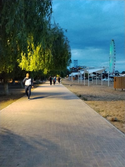 A group of people are walking along a paved pathway in what appears to be an amusement park or recreational area. The pathway is illuminated by artificial lights, casting shadows from the trees lining the left side of the path. The trees are lush and green, with some branches illuminated by yellow light. In the background, a Ferris wheel with green lights and water slides are visible, suggesting the location is an amusement park. Several white structures with triangular canopies are set up on a sandy area to the right of the path.

The sky is a deep blue, indicating it is either dusk or dawn. The overall atmosphere is calm and relaxed, with people enjoying an evening stroll. The subjects are casually dressed, and their body language suggests a leisurely activity. The scene is well-lit, creating a pleasant ambiance for an evening outing. The location appears to be near a body of water, possibly a beach, given the sandy area. There is no visible text in the image.
