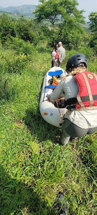 The image depicts an outdoor scene in a vegetated, hilly area of Kikagati, Uganda. In the foreground, a person is crouched over a white inflatable raft, viewed from behind. This individual wears a black helmet, a light grey long-sleeved shirt, dark grey pants, and a red personal flotation device (PFD). Their hands are positioned near the top edge of the raft, which rests on dense green grass. In the mid-ground, two other individuals stand on the grass behind the raft. The first, closer to the raft, has dark skin and wears a light-colored shirt and a red PFD, looking towards the foreground. Behind them, a person with lighter skin wears a light-colored long-sleeved shirt and a light-colored baseball cap, also looking forward. The background features abundant green bushes and trees, transitioning into distant rolling hills under a bright, clear sky. The scene suggests preparation or activity involving the inflatable raft in a natural, rural setting.