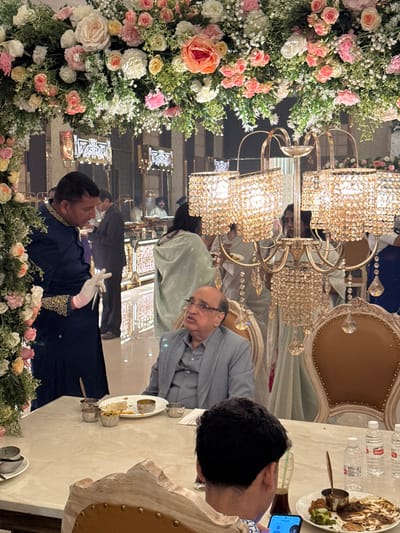 An indoor event with floral arrangements, chandeliers, and tables set for dining. People are seated at tables, including an older man in glasses and a younger individual using a cell phone. Another person wears white gloves and a dark-colored garment with gold trim. The scene is reflected in a mirrored wall, creating a repeated visual effect. Water bottles and plates of food are visible on the tables.