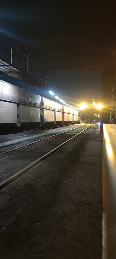The image depicts a freight train at a station at night. The train consists of multiple grey cars with visible numbering on some of them. The train is parked on a railway track, with another set of tracks to its right. The surrounding ground is dirt and gravel. Overhead, there is a structure with lights, illuminating the scene. In the background, there are more lights, some of which appear to be streetlights or station lighting, and a few distant vehicles are visible. The overall impression is that of an industrial or transportation hub at rest during nighttime hours. This scene is not specifically identifiable to Salzgitter, Germany without further contextual information.