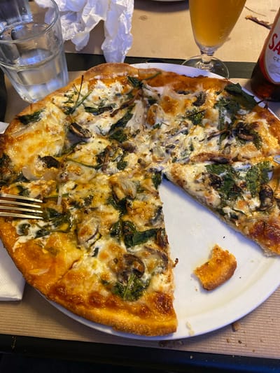 A whole pizza with a slice removed is on a white plate, on a table. The pizza is topped with cheese, mushrooms, and spinach. To the left of the pizza is a glass of water. To the right of the pizza is a glass of beer and a bottle of beer. The image was taken at a restaurant in Olhão, Portugal.