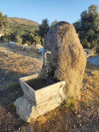 The image depicts an outdoor scene in Ledrada, Spain, featuring a natural water source. A large, rough-textured boulder serves as the backdrop for a functioning tap, from which water is flowing into a concrete trough situated below. The trough appears to be partially filled with water. The surrounding environment is arid, with dry grass, small stones, and sparse vegetation covering the ground. In the background, a hillside is visible, dotted with trees and a few wind turbines, suggesting a rural or semi-rural setting. The sky is clear and blue, indicating fair weather. The lighting suggests it is either early morning or late afternoon. There are no people or animals visible in the frame.