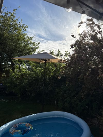 The scene is in a backyard, likely in a residential area of Hazlemere, United Kingdom, given the mention of the location. An inflatable blue swimming pool occupies the right side of the frame, containing water and a blue and yellow inflatable ring. Adjacent to the pool is dense greenery, including trees and bushes. A beige umbrella is positioned amidst the foliage, providing shade. The sky above is visible, featuring a mix of blue sky and scattered clouds. In the upper left corner, string lights are hanging, partially obscured. The scene captures a sunny outdoor setting in a private garden.