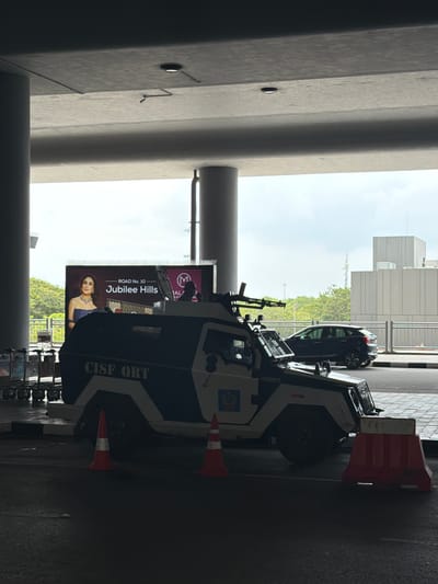 The image depicts a security scene likely at an airport or a similar high-security area in Hyderabad, India. A dark-colored armored vehicle, identified by the "CISF QRT" (Quick Response Team) marking on its side, is parked. The vehicle is equipped with what appears to be a mounted weapon system. In the background, a large advertisement featuring a woman and text including "ROAD No. 10 Jubilee Hills" is visible. Jubilee Hills is a prominent locality in Hyderabad known for its residential and commercial establishments. A dark-colored sedan is parked on the road behind the armored vehicle. Orange traffic cones and a red and white traffic barrier are also present on the ground. The scene is situated under a concrete overpass or structure, with portions of airport trolleys visible on the left. The sky is overcast.