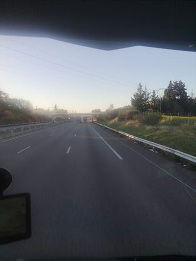 The image depicts a forensic view from inside a vehicle traveling on a multi-lane highway in Sauzet, France, under a clear, bright sky with low sunlight suggesting early morning or late afternoon. The road features two lanes in the direction of travel, marked by white broken and solid lines, and is paved with asphalt. Concrete median barriers separate the opposing direction of traffic on the left, while a white metal guardrail borders the right shoulder. In the distance, several large vehicles, likely trucks, are visible on the highway, approaching a light-colored concrete bridge or overpass that spans the road. Overhead, multiple parallel power lines traverse the frame against the sky. The highway is flanked by grassy verges, dense green bushes, and various trees on both sides. On the far right, beyond the guardrail, there is a field containing yellow flowering vegetation. A dark object, possibly a GPS device or camera mount, is partially visible in the lower-left corner of the foreground. No individuals are discernible.
