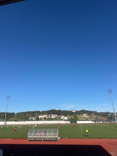 A bright blue sky fills the majority of the image. A stadium with a green soccer field bordered by a red track is visible on the right side. Soccer players wearing red uniforms are scattered across the field. A building with an orange tower and a white building are visible in the distance, above the top side of the field and the town. A row of trees line the background above the stadium. Lighting poles are on the near and far side of the field. The scene appears to be a soccer game in progress. The image features a soccer field along the Av. da Noruega, Valpaços, Portugal, based on the tower and solar panels on the horizon.