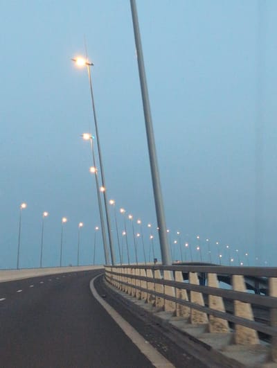 The image shows a view from a moving vehicle on a bridge at dusk or dawn. The sky is a pale blue, and streetlights are illuminated along the bridge. The road is asphalt with white lane markings. A concrete barrier with metal railings lines the right side of the bridge. In the background, the structures of the bridge are visible, along with more streetlights. The location is identified as the city of Oudalle, France. There are no people visible in the image.