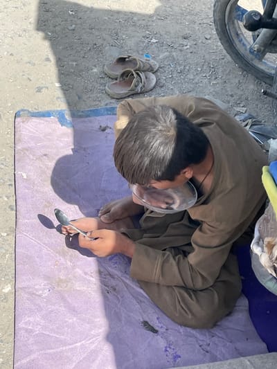 A single male individual is seated on a crumpled, light purple mat on an unpaved dirt and gravel surface. He wears a loose-fitting, brown or olive-colored garment and is barefoot. His head is bowed, with a clear, shallow, circular plastic object positioned over his lower face, obscuring his mouth and nose. His right hand holds a silver spoon, oriented with the bowl facing downwards towards the mat. His left hand rests near his right knee. A pair of worn, brown open-toed sandals are on the ground behind the individual. In the upper right background, the lower portion of a blue motorcycle wheel with spokes is partially visible. Strong shadows are cast across the mat and ground, indicating direct sunlight. Crumpled white and yellow/green material is visible to the individual's right, within a blue container or bag. The scene is located in the city of Spin Boldak, Afghanistan.