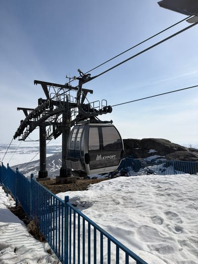 The image displays a ski lift gondola system located in a mountainous, snow-covered environment, identified as Zelenaya Polyana, Russia. A large, dark-grey metal pylon, part of the ski lift structure, stands prominently in the mid-ground, supported by a concrete base. Multiple heavy-duty steel cables extend from the pylon, supporting a modern, silver-grey and black gondola cabin. The cabin is numbered "17" and features the Cyrillic text "КУРОРТ" (Resort) along with a stylized mountain logo on its side, beneath tinted windows.
In the immediate foreground, a blue metal safety fence, composed of vertical railings, runs horizontally across the lower portion of the frame. The ground directly beneath and around the fence is covered with patches of snow, interspersed with areas of exposed dark earth and rock, suggesting melting conditions. Further to the right of the gondola, large, dark rock outcrops are visible.
The background presents a vast panoramic view of a distant landscape, characterized by snow-covered hills and mountains with sparse, bare vegetation. A large, possibly frozen, body of water is visible in the far left distance. The sky above is clear and light blue, indicating fair weather conditions. No individuals are present in the scene.
