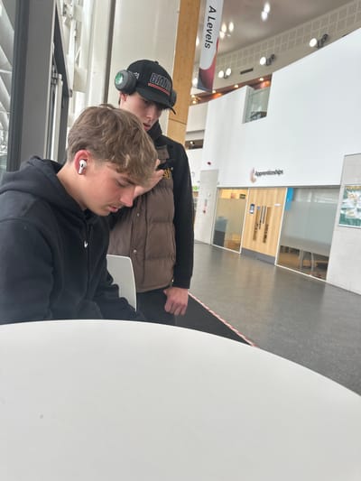 The image shows two males sitting at a white table in a brightly lit interior space. The individual closest to the camera is wearing a black hoodie and a white earbud in his right ear. Behind him, another male is wearing a dark cap with "GIANTS" embroidered on it, a black jacket, headphones around his neck, and a brown puffy vest. The environment is modern, with large windows and a sign in the background that reads "A Levels". Further back, the signs indicating "Apprenticeships" can be observed. The location is in Wistaston, United Kingdom.