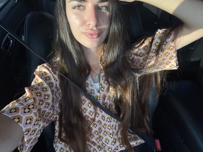 A young woman with long, dark hair and light-colored eyes is seated in the driver's seat of a car. She is wearing a collared, short-sleeved shirt with a geometric pattern in shades of pink, orange, yellow, and black. A silver necklace with a small pendant is visible around her neck. The car's interior is dark, with black leather seats and a seatbelt diagonally across her chest. Sunlight illuminates her face and hair, casting shadows. The scene appears to be in Deerfield Beach, Florida, United States of America.