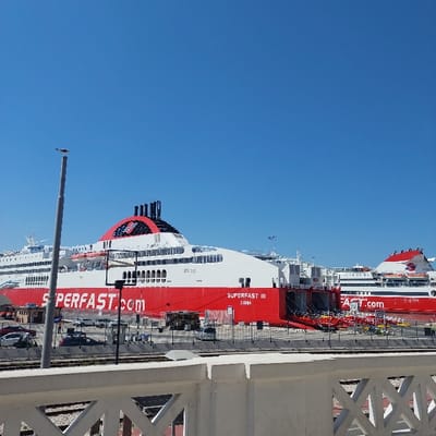 The image presents a daytime scene at the Port of Ancona, Italy. The view is dominated by two large, red-hulled, white-superstructured passenger and vehicle ferries docked side-by-side under a clear, bright blue sky.

The primary vessel, positioned centrally, is identified as "SUPERFAST III XANIA" with "SUPERFAST.com" prominently displayed in white lettering on its red hull. This ferry features multiple black funnels with a red and white logo and appears to have stern ramps open or prepared for vehicle loading/unloading. A second, similar-looking ferry, also red and white with a visible ".com" branding, is partially seen docked to the right of the primary vessel.

In the immediate foreground, a white concrete balustrade with an open, repeating geometric pattern is visible, suggesting an elevated viewing position such as a balcony or pedestrian walkway. Below this, parallel railway tracks traverse a paved area. Between the tracks and the docked ferries, the port's dockside is visible, populated with various parked passenger vehicles, including cars and vans. Ancillary port infrastructure, such as tall light poles, shorter lampposts, security fencing, and small modular buildings or kiosks, are dispersed throughout the dock area. No distinct individuals or specific actions are discernible, with the focus on the vessels and the port environment itself.