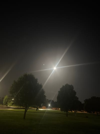 The image captures a nighttime scene in Columbia, Missouri, United States of America. A bright, luminous moon, appearing as a distinct circle, dominates the sky, creating a significant lens flare that extends across the frame in a cross-like pattern. The moon is partially obscured by a hazy atmosphere, which also contributes to the overall glow. Several trees are visible in the foreground and midground, silhouetted against the moonlit sky. The ground is covered in grass, suggesting an outdoor park or open area. In the background, there are hints of man-made structures, including streetlights and possibly buildings, illuminated by artificial lights. The scene is serene and devoid of any human presence.