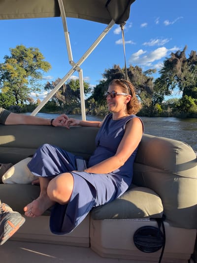 A woman in a blue dress and sunglasses sits on an olive-green pontoon boat. She is smiling and appears to be holding hands with someone whose arm is visible. The setting appears to be a river or waterway lined with trees, possibly with Spanish moss hanging from them. A silver-colored boat canopy frame is visible overhead, with a blue sky and scattered clouds in the background. The scene gives the impression of a leisurely boat ride. The city in question is not visible in the picture.