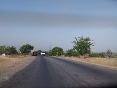 The image shows a paved road in a rural area of Lakweme, Nigeria, located at 9.707 latitude and 11.432 longitude. The road is empty except for two vehicles travelling in the same direction. The first vehicle is a large truck carrying a load of goods, and the second vehicle is a white pick-up truck. Both vehicles are moving from left to right in the image. The road is bordered by trees and shrubs on both sides. The sky is clear and blue, and the sun is shining brightly. The ground is dry and dusty, and there is some evidence of vegetation. The ground is brown and dusty, and there are some trees and shrubs along the side of the road. The sky is a pale blue, and there are no clouds visible.