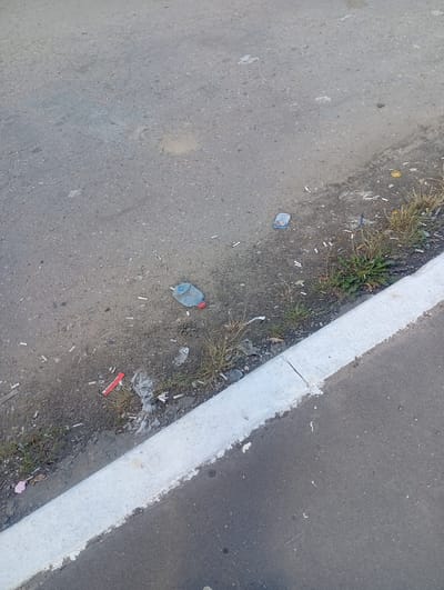 The image shows a roadside scene in Oktyabrskiy, Russia. The foreground features a portion of a paved road with a white concrete curb. Scattered along the edge of the road and on the road surface itself is a significant amount of litter, primarily cigarette butts. There is also a discarded plastic bottle with a red cap, a small blue package, and some crumpled plastic trash. Patches of dry grass and weeds grow along the curb. The road surface appears to be asphalt, with some small stones and debris visible. The overall impression is one of neglect and urban pollution. The lighting suggests it is daytime, with no specific indication of weather conditions. No people are visible in the image.