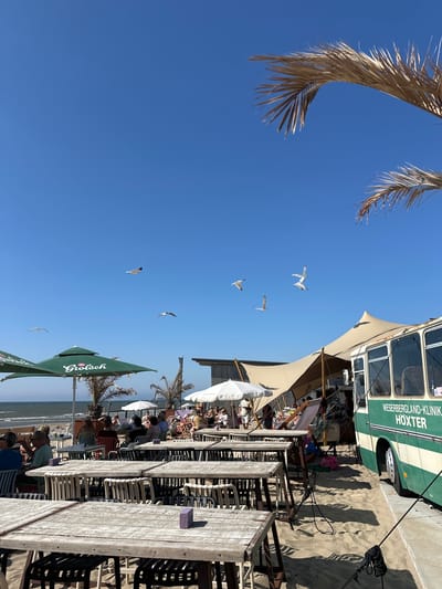 The image depicts a lively beachside scene at a cafe or restaurant in Zandvoort, Netherlands. In the foreground, several weathered wooden tables and chairs are set up on the sand, indicating an outdoor dining area. To the left, two large green umbrellas with the "Grolsch" logo provide shade. Beyond the tables, people are seated and enjoying themselves, some under additional white umbrellas.
A prominent feature of the scene is a vintage green and white bus, partially covered by a large, light-colored tent structure. The bus has text on its side reading "WESERBERGLAND-KLINIK HOXTER." The overall ambiance suggests a popular spot for relaxation and socializing, likely on a sunny day given the clear blue sky and the presence of beachgoers. Seagulls are visible flying in the sky, adding to the coastal atmosphere. The background shows the ocean and a sandy beach, characteristic of Zandvoort's coastal landscape.