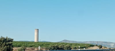 The image captures a landscape scene under a clear blue sky. Dominating the mid-ground is a tall, cylindrical concrete structure, possibly a water tower, with a weathered appearance. It is situated behind a dense field of green foliage, likely crops or vineyards. To the left of this structure, a large, leafy green tree stands prominently. In the far distance, rolling hills and mountains form the horizon, with a peak that appears to have some structure on it. The foreground shows a road or highway barrier, with some signage visible. The overall environment suggests a rural or semi-rural setting, possibly on the outskirts of a town. The specific location is identified as Belvís de Monroy, Spain, implying this is a view of the Spanish countryside. There are no people or animals visible in the frame.