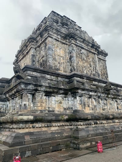 The image shows the exterior of a large stone temple, likely a Buddhist temple, located in Mungkid, Indonesia, at the coordinates -7.605 latitude and 110.23 longitude. The temple is made of weathered gray stone blocks and is covered in moss and lichen. There are several decorative carvings on the temple walls. The temple is surrounded by a paved courtyard, with a pink propane tank in the foreground. The sky is overcast.