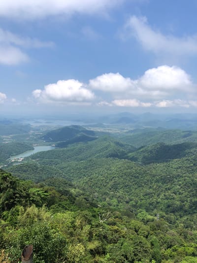 The image is a high-angle view of a lush green mountainous landscape with a body of water in the middle ground. The sky is blue with scattered white clouds. The foreground shows dense vegetation with some rocky elements. In the distance, multiple layers of rolling hills recede into a hazy horizon. A portion of a metallic structure, possibly a pipe or a railing, is visible in the lower left corner of the frame. The scene appears to be a natural landscape, likely a viewpoint overlooking a valley or reservoir, characteristic of the scenic mountainous terrain found in areas such as Nam Hải, Vietnam, known for its natural beauty and elevated vistas.