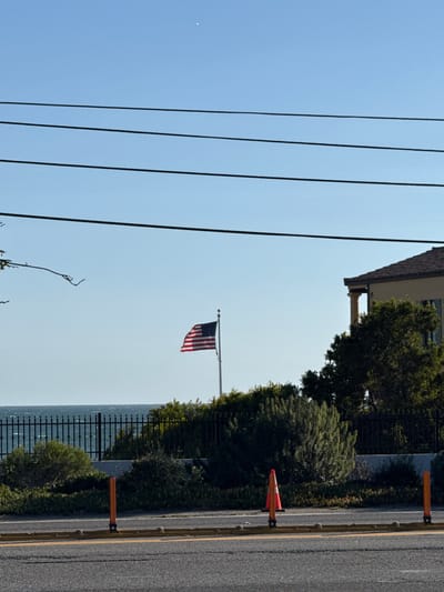 In Malibu, California, an American flag flies on a flagpole against a clear blue sky. The flag is positioned near a dark metal fence that borders a body of water, likely the Pacific Ocean. Behind the fence, lush green bushes and trees are visible, with a portion of a building with a beige facade and a brown tiled roof appearing in the upper right of the frame. In the foreground, a paved road with yellow lane markings is present, with orange traffic cones and bollards placed along the side, suggesting ongoing construction or maintenance. Power lines are strung across the upper portion of the image.