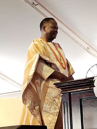 The image captures a dark-skinned man, likely middle-aged or older, standing with a pleasant, subtle smile, looking towards his right. He is dressed in traditional Nigerian attire, a flowing Agbada, made of a light beige or golden fabric. The garment features intricate patterns, including lighter floral or abstract motifs and distinct vertical stripes. Around his neck, he wears multiple strands of vibrant red coral beads, a common cultural accessory in Nigerian ceremonial dress.
He stands behind a dark wooden podium or lectern, visible in the lower right portion of the frame, which has decorative horizontal ridges and vertical paneling. A white object, possibly a floral arrangement or decoration, is partially visible next to the podium on the right. A faint mesh structure, possibly part of a fan, can be seen further to the right.
The setting appears to be indoors or under a large tent, characterized by a light, off-white draped fabric ceiling with visible creases. Some dark fixtures or wires are attached to the ceiling structure in the upper left. The overall ambiance suggests a formal or ceremonial occasion. The man's attire and accessories strongly indicate a cultural event taking place in Nigeria, aligning with the provided location context of Dushepe. His serene expression suggests he is engaged in or observing the proceedings of the event. No visible text is present in the image.