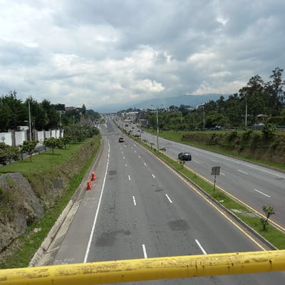 A multi-lane highway stretches into the distance under a cloudy sky. Vehicles are sparsely scattered along the roadway. Greenery borders both sides of the highway, with buildings visible in the background. A yellow structure occupies the right foreground. The highway is marked with white lines and a yellow divider line. A mountain range is faintly visible on the horizon.