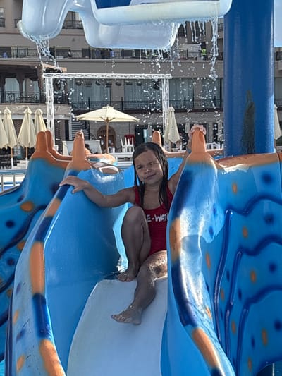 A young girl in a red swimsuit is positioned on a blue and orange water slide. The slide is part of a water park facility in the city of Al Ahia', Egypt. Water sprays from a structure nearby. In the background, buildings with balconies and awnings are visible, as are several white sun umbrellas.