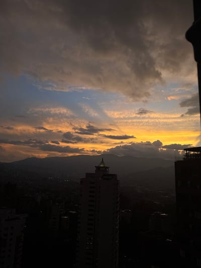 The scene captures an urban panorama at dusk in Medellín, Colombia, from an elevated perspective. The sky exhibits a sunset with stratified cloud cover: dark grey cumuliform clouds in the upper left, transitioning to a narrow horizontal band of blue, then expanding into a broad expanse of orange and yellow light towards the right and horizon. Silhouetted mountains define the distant skyline. The city of Medellín occupies the lower third of the image, rendered in deep shadow, with numerous structures indistinct against the twilight. A prominent, light-toned high-rise building featuring a conical, metallic-capped roof is centrally positioned in the mid-ground. Additional dark building masses are visible in the foreground, including a structure on the far right displaying rooftop foliage. No human subjects are present. A dark, vertical structural element partially obstructs the extreme right edge of the frame.