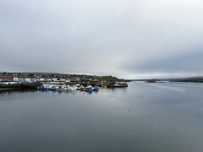This is an image of a harbor in Portmagee, Ireland, under a cloudy sky. The scene depicts a row of colorful buildings lining the waterfront, with a dock and several boats moored in the calm water. The boats are a mix of white and blue, with some fishing vessels also visible. Beyond the harbor, a grassy hillside rises, dotted with more houses and buildings. In the distance, islands can be seen in the sea, shrouded in mist. The overall impression is one of a quiet, working fishing village on a gray, overcast day. The water reflects the sky, creating a muted, serene atmosphere. There are no people visible in the image, and the activity is limited to the boats at rest in the harbor. The time of day is unclear due to the overcast conditions, but the lighting suggests it is daytime. Some text is visible on a sign near the harbor: "St. Brendan's Restaurant".
