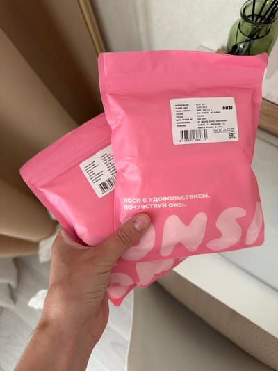 A person is holding two pink resealable bags with a white label on the front. The bags appear to contain socks. The label on the visible bag shows product information including "SLIM TOP", "S(44) MILK", "92% ХЛОПОК, 8% ЛАЙКРА", "РОССИЯ", "Май: 2026", and an address in Cheboksary. The bags have the brand name "ONSI" printed in large white letters, and a slogan in Russian that translates to "Wear with pleasure. Feel ONSI." The background is a blurred interior scene with a light-colored wall, a wooden shelf, and a green glass bottle with dark reeds. The flooring is partially visible as light-colored laminate. The overall scene is indoors and well-lit. No specific location in the city of Semiluki, Russia, is depicted or indicated in the image.