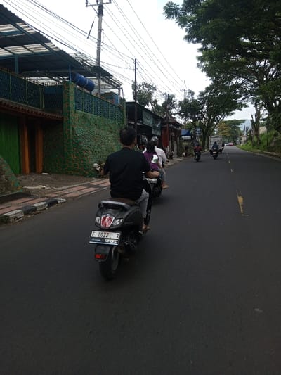 The image depicts a daytime street scene, with multiple individuals traveling on scooters and motorcycles. The primary view is from behind a dark-colored scooter, identified by its license plate "F 2022 X Sumare 04-29," indicating a location in Indonesia, specifically the Bogor region (F prefix), rather than Khajuripada, India.
In the immediate foreground, a male individual wearing a dark short-sleeved shirt and light-colored pants, with bare feet, operates a dark scooter. Directly behind him, another scooter is visible, carrying at least one passenger in a purple top. Further down the asphalt road, several other motorcycles or scooters are observed moving in the same direction, away from the camera. A white truck and other distant vehicles are also present on the road.
To the left of the road, a sidewalk with a black and white striped curb borders a green-patterned stone retaining wall or fence. Behind this wall, structures with an orange wall segment and a green door are visible. Above these structures, a multi-layered roof system with what appears to be blue cylindrical water tanks is present. Numerous overhead power lines traverse the scene, supported by utility poles.
The right side of the road is lined with mature trees featuring dense green foliage. In the distance, hills or mountains are discernible. The sky is overcast with a uniform light grey cloud cover. The road itself is asphalt, featuring a broken yellow line on its surface, indicating traffic lanes.