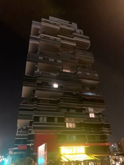 The image depicts a nighttime urban scene centered on a tall, modern building with a distinctive, multi-tiered, and cantilevered architectural design. The building, located at 2 Rue des Péniches, Issy-les-Moulineaux, France, appears to be the Tour des Péniches (also known as Arboretum) complex.

The structure is predominantly dark grey with lighter grey or white banding along its numerous projecting balconies and terraces, creating a stepped or block-like facade. A large, rectangular section of the lower building facade is colored bright red. Multiple windows are visible across the building's height, with several illuminated from within, including a distinctively bright, square light on a mid-level floor towards the left side of the frame.

At the ground level, a commercial establishment is visible at the base of the building. It features a bright yellow awning and a prominent, rectangular yellow sign that clearly displays the name "PAULETTE" in dark lettering. Undistinguishable human figures are present beneath the awning, suggesting pedestrian activity. To the far left, a portion of a vertical sign or banner with blue and white elements is partially obscured. The sky is dark, indicative of nighttime conditions, and scattered, blurred artificial lights from other street elements are visible at the bottom edges of the frame.