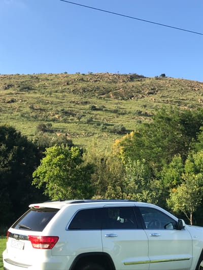 A white SUV parks adjacent to a green hillside under a clear blue sky. Sparse vegetation covers the hillside, with trees and shrubs adding dimension. A utility wire extends across the upper left of the frame. The vehicle's rear taillight, tinted windows, and door handles are visible, along with the side mirror and a glimpse of the front wheel.