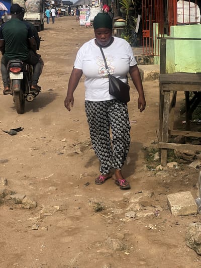 This daytime image, captured in Ijegun, Nigeria, features a woman as the main subject, standing on a rough dirt ground. She is facing slightly towards the viewer with a neutral expression, wearing a white short-sleeved t-shirt adorned with a cartoon graphic of rabbits and partial text that reads "I'M R...". She also wears black and white patterned trousers, a dark green head covering, a black shoulder bag, and pink flip-flop sandals.

The ground beneath her and extending throughout the foreground is unpaved, composed of dry, light brown dirt with scattered small rocks and debris. In the mid-ground to the left, a person is visible on a black motorcycle, its license plate partially visible with "RC" or "8C" followed by numbers/letters. The background shows a bustling street scene with several blurred figures, suggesting other people moving about. Structures in the background include simple buildings with light green and grey walls, some with visible rebar or metal railings, and a prominent wooden scaffolding-like structure behind the main subject.

Visible text on signs in the background includes "NO SUPER STORES" and "POS Services," indicating a commercial or service-oriented area. The bright, even lighting suggests it is a clear day. The overall impression is that of an active, everyday street or market environment in a local community.