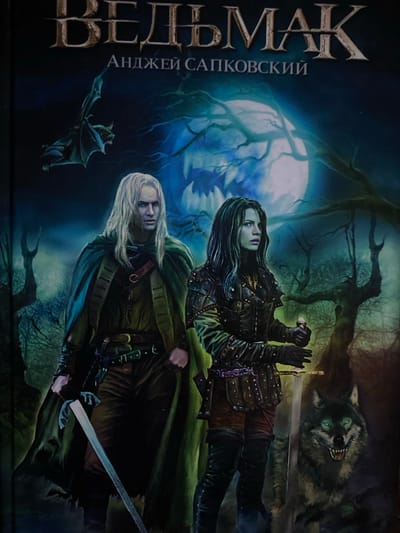 This book cover depicts a dark fantasy scene set in a moonlit forest at night. In the foreground, a man with long, white hair, a pale face, and a determined expression stands, clad in a dark green cloak and dark clothing. He holds a silver-bladed sword pointing downwards in his right hand. Behind him, a woman with long, dark hair and an intense gaze is visible, wearing dark, studded leather armor. She clutches a short sword or dagger, held upwards.

The background features gnarled, bare trees silhouetted against a large, luminous full moon that casts a blue-white glow on the scene. In the upper right, a snarling wolf with glowing blue eyes and bared teeth peers from the shadows. A small, dark, bat-like creature flies in the lower left corner. The overall mood is mysterious and tense, suggesting an impending encounter or vigilance in a dangerous environment.

Visible text in Cyrillic along the left side of the cover reads "ВЕДЬМАК" (The Witcher) in large letters, with "АНДЖЕЙ САПКОВСКИЙ" (Andrzej Sapkowski) below it, identifying the book and its author.