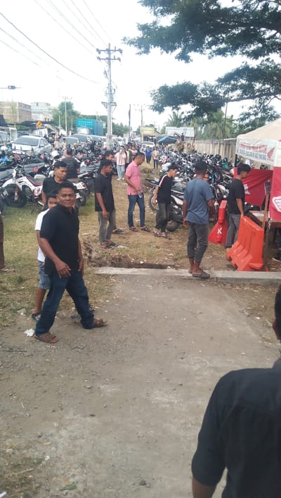 The image depicts an outdoor scene in Keude Trienggadeng, Indonesia, likely a parking area or event space. In the foreground, a man in a black polo shirt and blue jeans, wearing brown sandals, is walking towards the camera. Behind him, a child in a white t-shirt and denim shorts is also walking. To the left, a white car is parked amongst numerous motorcycles. The background is filled with a dense collection of parked motorcycles, suggesting a crowded event or popular gathering place. Several people, predominantly men, are visible among the motorcycles, some standing and observing, while others appear to be walking or interacting. The environment includes power lines and poles, a traffic light, buildings in the distance, and trees. There are also orange traffic barriers and a tent structure with a sign, possibly indicating an event or information booth. The ground is a mix of gravel and dirt.