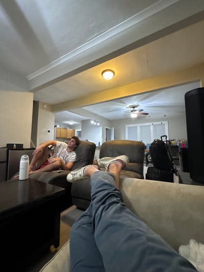 The scene depicts an indoor residential common area, identified as being located in South Bend, United States.
**People:**
* A male individual with light skin and dark hair is visible in the midground, reclining on a dark brown, armchaired sofa. He wears a white t-shirt with a graphic design and red shorts. His right arm is bent, resting on his leg, and his gaze is directed towards the right side of the frame.
* The lower legs of a second individual are present in the immediate foreground, positioned with feet propped up on a light-colored sofa or ottoman. This individual wears blue-grey trousers and white sneakers with red accents.
**Objects:**
* A dark wooden coffee table occupies the foreground, displaying a white insulated water bottle.
* A small, dark rectangular object (possibly a phone or remote) rests on the armrest of the sofa next to the reclining male.
* Behind the reclining male, another dark brown recliner-style chair is visible.
* Further back, a black backpack with a telescopic handle is stacked on top of a smaller black bag.
* A large, black, textured speaker or similar appliance is partially visible along the far right edge of the frame.
* The background reveals an open-concept kitchen area with light wood upper cabinets and a partial view of stainless steel appliances, including a refrigerator on the left.
* A circular white thermostat or control device is mounted on the wall to the left of the kitchen entrance.
* Overhead, the ceiling features decorative beams or molding, along with multiple artificial light sources, including a flush-mount fixture, recessed lights, and a central ceiling fan with an integrated light kit.
* Windows with white blinds or coverings are visible in the distant background.
**Actions/Interactions:**
* The male in the midground is reclined in a relaxed posture.
* The individual in the foreground has their legs propped up, indicating a relaxed state.
* No direct interaction between the individuals is depicted.
**Environment:**
The space is a well-lit, open-concept interior with light-colored, possibly beige or off-white, walls. The ceiling has a textured appearance. Artificial lighting is active throughout the area. The overall setting is casual and domestic.