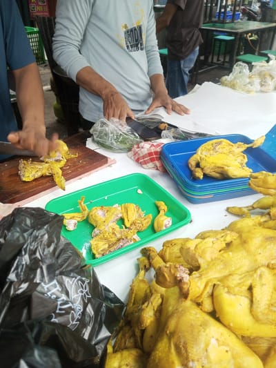 The image depicts a bustling outdoor food stall in Houaythat, Laos, specializing in prepared chicken. In the foreground, a person wearing a dark blue short-sleeved shirt is actively cutting a cooked, yellow-hued chicken on a rectangular wooden cutting board with a large knife. Partially dismembered chicken pieces are visible on the board. Adjacent to this, a bright green plastic tray holds additional cut chicken portions. A stack of blue plastic trays contains another whole cooked chicken. A large pile of multiple yellowish-cooked chickens dominates the right foreground, suggesting a bulk display for sale.
In the mid-ground, a second individual wearing a light blue long-sleeved shirt with a yellow graphic has their hands resting on white paper laid across a surface, with a black smartphone and a clear plastic bag containing green leafy vegetables nearby. A red and white checkered cloth is also present. In the background, a third person, dressed in a brown shirt and blue jeans, is partially visible near a dark metal table equipped with green stools. Further background elements include portions of a dark-colored motorcycle, a green plastic basket, and a red sign marked "POS 55." A black plastic bag with a white floral design partially obscures the lower left foreground. The scene is illuminated by what appears to be natural light, indicating an open-air or semi-covered market environment focused on food preparation and retail.
